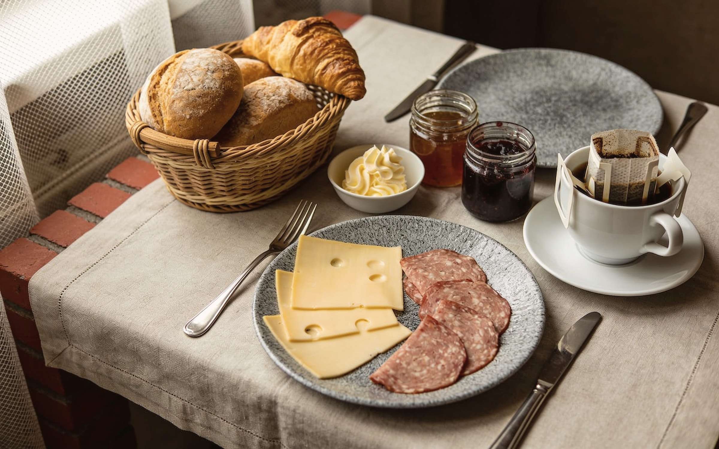 Frühstückstisch am Fenster: Brötchenkorb, Käse und Aufschnitt auf Steingut, Butter, Marmelade und eine Tasse Kaffee im Morgenlicht